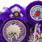 A man wearing a large, elaborate, feather Mardi Gras Indian costume. He's holding an equally elaborate walking stick